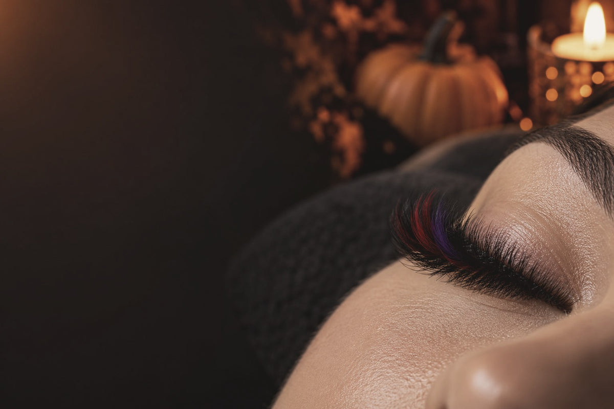 Closed eye with black volume lash extensions and red and purple colour lashes, with pumpkins and candlelight behind